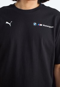 Mustan puuvillan T-paita, jossa on valkoinen Puma-logo ja BMW Motorsport -teksti pyöreällä logolla, lyhyet hihat ja klassinen malli.