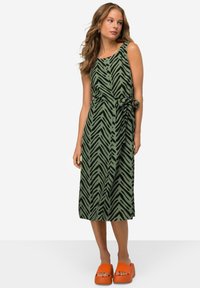 Grünes Kleid mit schwarzem Zickzackmuster, knielang, ärmellos, tailliert mit Bindeband, kombiniert mit orangen Plattform-Sandalen.