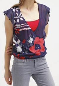 Genomskinlig navyblå blus med korta fjärilsärmar, med färgglada blommönster i rött, vitt och rosa. V-ringning. Kombinerad med gråjeans.