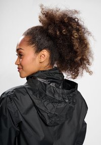 Kobieta z kręconymi włosami związanymi do tyłu, ubrana w czarną wiatrówkę z kapturem, pokazana z profilu na jednolitym jasnym tle.