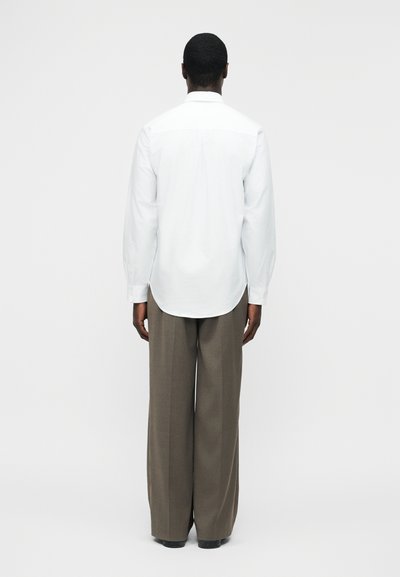 Άνδρας που κοιτά προς τα πίσω, φοράει λευκό μακρυμάνικο πουκάμισο και φαρδιά καφέ παντελόνια με φαρδύ πατρόν, πάνω σε απλό ανοιχτόχρωμο φόντο.