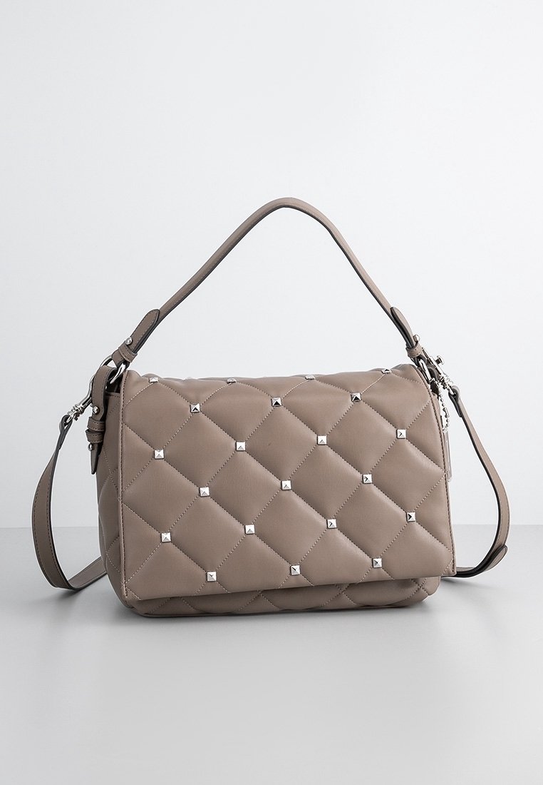Quiltet beige skuldertaske med sølvnitter, der har et top håndtag og en justerbar skulderrem. Blødt materiale med en tekstureret overflade.