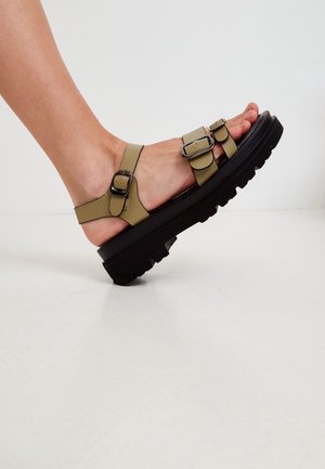 Sandales à sangle verte avec deux boucles ajustables, semelle noire épaisse avec texture antidérapante, design à bout ouvert et finition lisse.