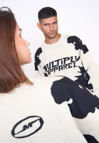 Sweatshirt de culoare crem cu imprimeu abstract de vacă în negru și textul "MULTIPLY APPAREL." Include un logo pe spate.