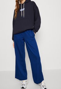 Námořnická mikina s kapucí s přední kapsou a světlými šňůrkami, doplněná širokými modrými kalhotami. Má volný střih a prodyšný materiál ze směsi bavlny.