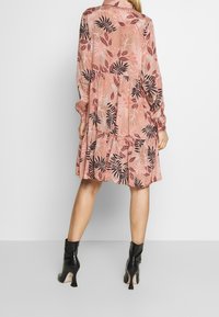 Robe rose pâle avec des manches longues, un col et une jupe évasée. Présente un motif de feuilles noires et est associée à des bottes à talons noires brillantes.