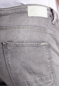Grå denimjeans har en bakficka, stickad detaljering och en vit läderlapp vid midjan. Tyget har en slät yta.