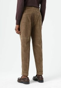 Bruine corduroy broek met een taps toelopende pasvorm, met achterzakken en opgerolde boorden, gecombineerd met donkergijze schoenen. Structuur van de stof zichtbaar.