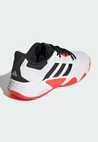 Valkoinen urheilukenkä mustilla yksityiskohdilla, jossa on kolme mustaa raitaa, punainen pohjan koristelu, verkkoyläosa ja pehmustettu kantapää tukea varten.