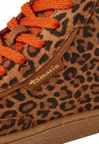 Gros plan sur une sneaker en daim marron avec un imprimé léopard noir et des lacets orange, mettant en avant le logo doré "Tamaris" sur le côté.