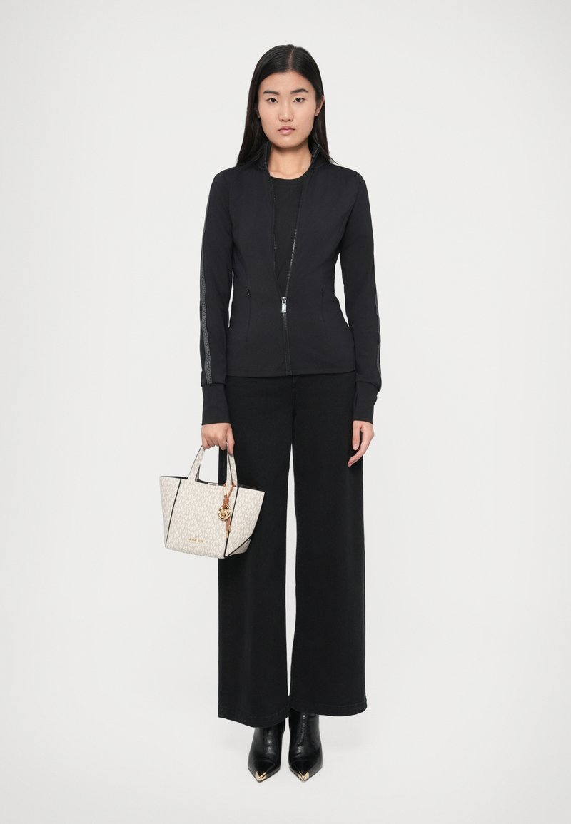 Veste noire zippée à manches longues, pantalon large noir, et petit sac à main blanc à motifs avec des accents dorés. Bottines noires à bout pointu.