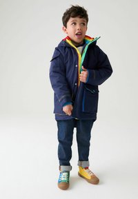 Mornarska vodoodporna jakna z barvitim notranjim podložjem, zelenimi in rumenimi zadrgami, v kombinaciji z jeans hlačami in večbarvnimi supergami.