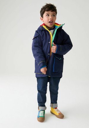 Giacca impermeabile blu con fodera colorata, cerniere verdi e gialle, abbinata a jeans in denim e sneaker multicolori.