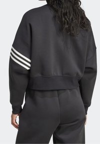 Persona che indossa una felpa nera a maniche corte con tre strisce bianche sulla manica sinistra e pantaloni neri abbinati, vista da dietro.