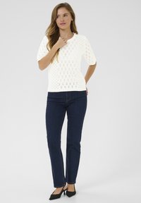 Top en tricot blanc avec un motif en losange, manches courtes, encolure ronde, associé à un jean droit bleu foncé et des escarpins noirs à bout pointu.