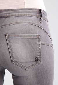 Šedé denimové džíny s texturovaným povrchem, se jednou zadní kapsou a vyšitým detailem poblíž rohu kapsy.