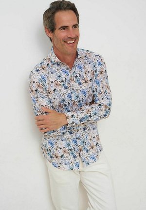 Mann lächelt mit verschränkten Armen, trägt ein langärmeliges Hemd mit blau-braunen Blumen und weiße Hose vor einem einfarbigen Hintergrund.