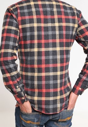 Uomo che indossa una camicia a quadri rossa, nera e beige a maniche lunghe con le mani nelle tasche, abbinata a jeans blu.