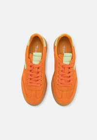 Paire de baskets en suède orange avec des accents vert clair et des lacets orange, vues de dessus sur un fond blanc.