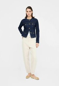 Kurtka dżinsowa w ciemnym niebieskim kolorze z złotymi guzikami, z okrągłym dekoltem, dopasowanym krojem i naszywanymi kieszeniami; zestawiona z kremowymi spodniami.