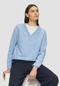 Hellblauer, gestrickter Pullover mit V-Ausschnitt. Mit gerippten Akzenten am Hals und an den Bündchen, getragen über einem weißen Hemd mit dunklen Hosen.
