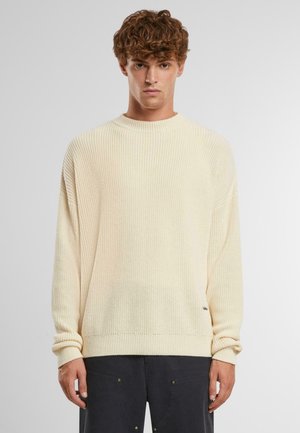 Licht beige gebreide trui met een geribbelde textuur, ronde halslijn en drop shoulders. Heeft opgerolde mouwen en een subtiele logo-detail.