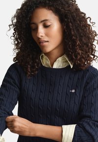 Granatowy sweter ze splotem warkoczowym wykonany z teksturowanej tkaniny, z prążkowanymi mankietami. Noszony na jasnółóżowej, paskowanej koszuli z kołnierzykiem i podwiniętymi rękawami.