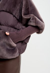 Hnědý kabát z umělého kožešinového materiálu, krátkého střihu, se hladkým prošívaným tělem, obsahující rukávy z jemného žebrovaného úpletu a měkkou texturu. Viditelné detaily kapsy na ruce.