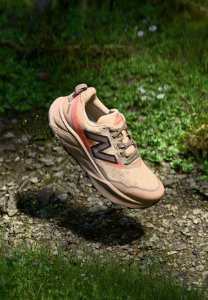 Hellbeiger New Balance Laufschuh mit orangen Akzenten, schwebend über einem Kiesweg, umgeben von grünem Gras und Pflanzen.