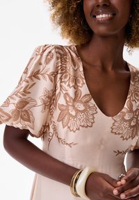 Lichtroze jurk met bruine bloemenborduring, korte puffmouwen en een diepe V-hals. Geaccessoireerd met armbanden en een ring.