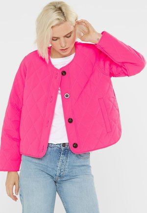 Blonde Frau passt ihre kurze, leuchtend pinkfarbene, gesteppte Jacke mit schwarzen Knöpfen über einem weißen Hemd und hellblauen Jeans vor einem weißen Hintergrund an.