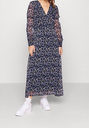 Mujer con un vestido largo de mangas largas y hasta el tobillo, transparente con estampado abstracto en azul y negro, combinado con calcetas blancas y zapatillas blancas.