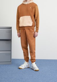 Felpa marrone con tasca beige chiaro e pantaloni jogger abbinati, entrambi realizzati in materiale morbido, abbinati a scarpe da ginnastica bianche e dorate. Hardware argentato visibile.