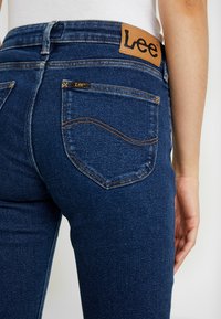 Mörkblå denimjeans med en ljusbrun läderetikett med texten "Lee" på baksidan av midjan. Märkbar sömnad och klassisk design med fem fickor.