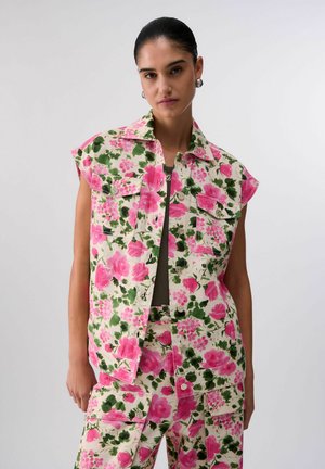 Donna che indossa una giacca senza maniche a fiori e pantaloni abbinati in tonalità rosa e verde con ampie tasche frontali, in piedi davanti a uno sfondo semplice.