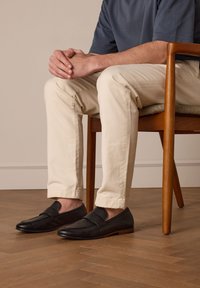 Zwarte leren instappers met een textuur op de bovenkant, platte zool en minimalistisch ontwerp. Draag met beige slim-fit broek, zittend op een houten stoel.
