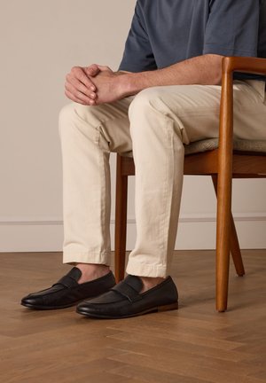 Zwarte leren instappers met een textuur op de bovenkant, platte zool en minimalistisch ontwerp. Draag met beige slim-fit broek, zittend op een houten stoel.
