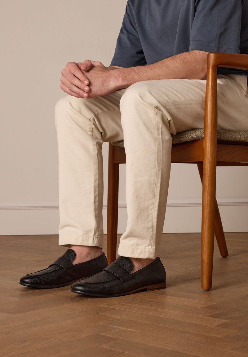 Zwarte leren instappers met een textuur op de bovenkant, platte zool en minimalistisch ontwerp. Draag met beige slim-fit broek, zittend op een houten stoel.