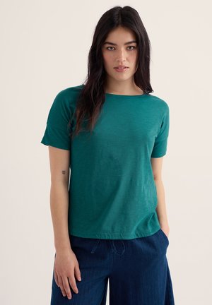 Jauna moteris su ilgais tamsiais plaukais, dėvinti žalsvai mėlyną trumpomis rankovėmis marškinėlį ir tamsiai mėlynas kelnes, stovi prieš šviesią neutralią foną.