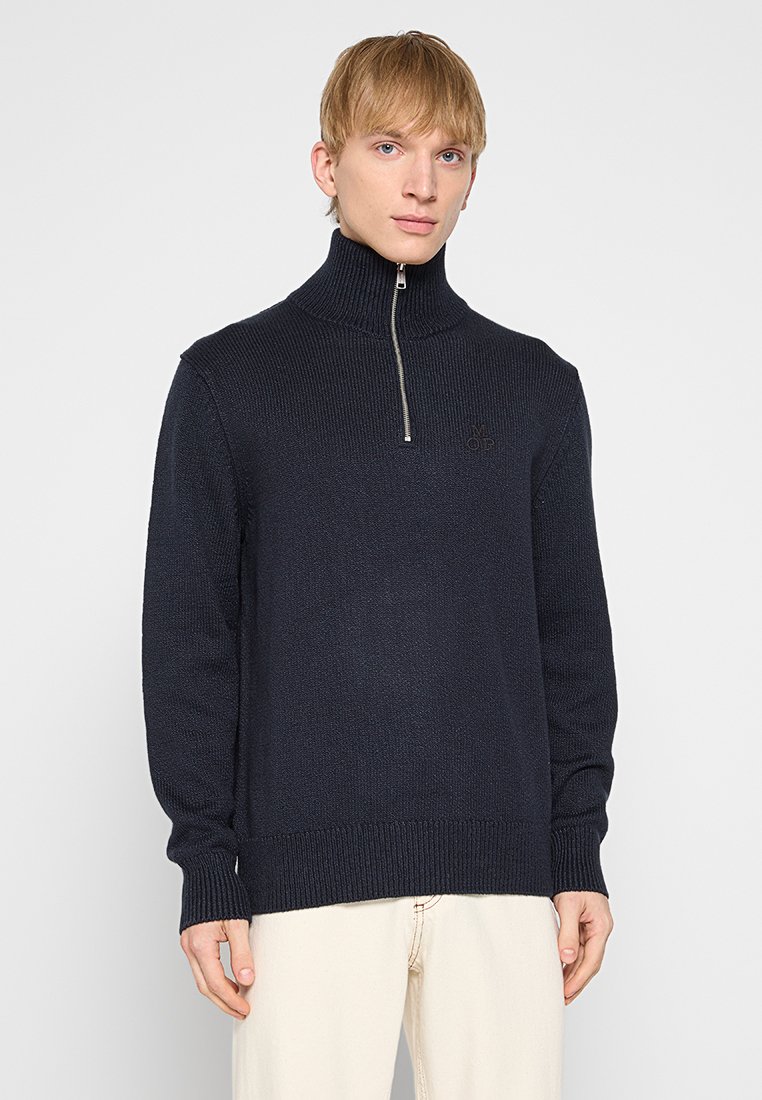 Granatowy sweter dziany z wysokim kołnierzem, pół zamkiem błyskawicznym oraz prążkowanymi mankietami i dołem. Posiada subtelną naszywkę z logo na klatce piersiowej.