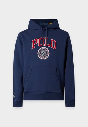 Giovane uomo che indossa una felpa con cappuccio Polo blu navy, pantaloni bianchi e mocassini beige, in piedi davanti a uno sfondo bianco uniforme.