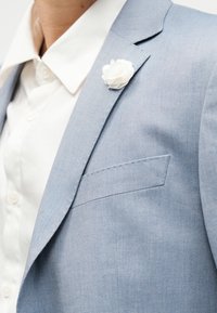 Άτομο με ανοιχτό γαλάζιο σακάκι, λευκό πουκάμισο και ένα μικρό λευκό λουλούδι καρφιτσωμένο στο πέτο του σακακιού.