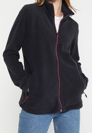 Kobieta ubrana w czarną polarkę z czerwonym zamkiem błyskawicznym i kieszeniami, w niebieskich dżinsach, stojąca z rękami w kieszeniach.