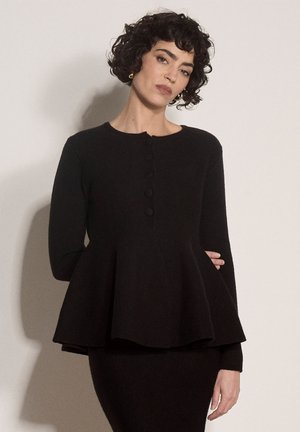 Woman with short curly hair wearing black long-sleeve peplum top with buttons and fitted skirt against plain light background.
