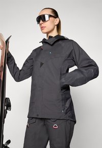 Black waterproof jacket with a hood, featuring mesh panels, a zip front, and adjustable cuffs. Paired with black ski pants and sunglasses.