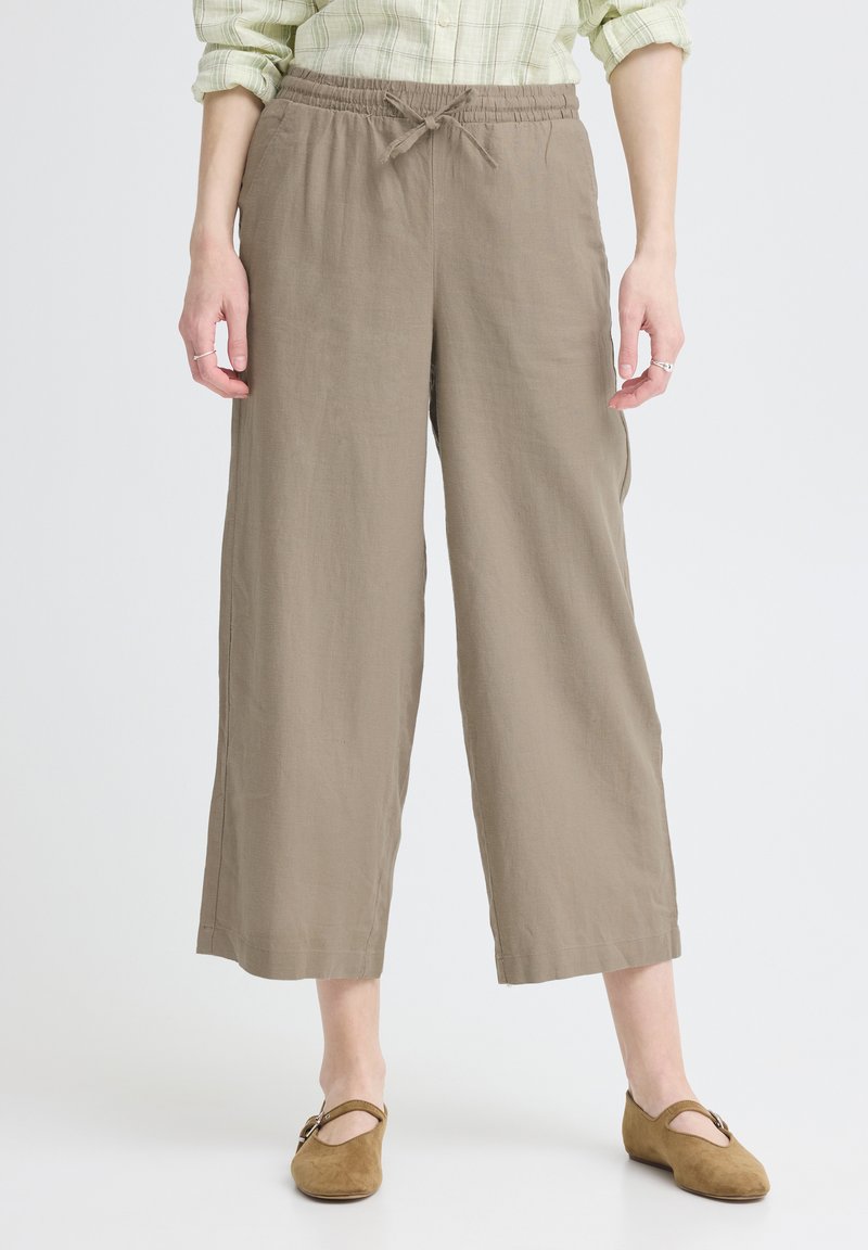 Femme portant un pantalon beige à jambes larges et courtes, avec une ceinture élastique et un cordon de serrage, associé à une chemise à carreaux vert clair et des chaussures Mary Jane marron.