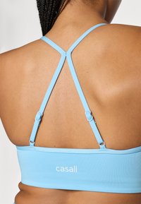 Αθλητικό σουτιέν σε ανοιχτό μπλε χρ ώμα με σχέδιο που διασταυρώνεται στην πλάτη, ρυθμιζόμενους ιμάντες και μικρό λευκό λογότυπο στη ζώνη. Λείο, ελαστικό ύφασμα.