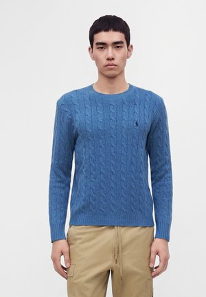 Jeune homme portant un pull torsadé bleu et un pantalon cargo beige, debout devant un fond blanc uni.