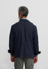 Chaqueta ligera de color azul marino con un cuello estructurado, epaulettes en los hombros y puños ajustables. Textura suave y diseño ajustado.