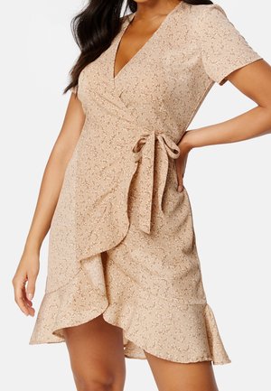 Mujer vistiendo un vestido cruzado beige con mangas cortas, pequeño estampado floral, bajo con volante y un lazo atado en la cintura.